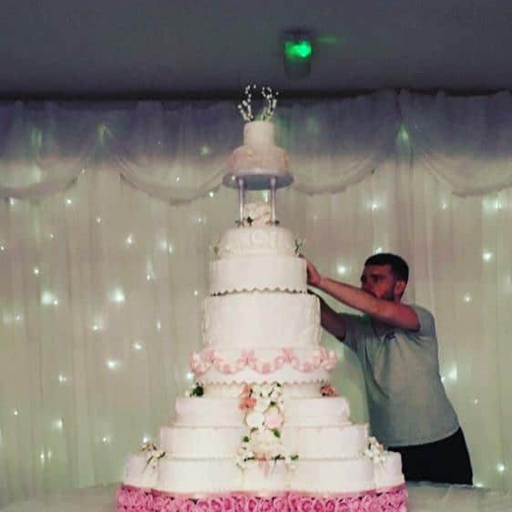 Extra large wedding cake pale pink and white adorned with sugar flowers and lace pieces separated with pillars on half noon stand and an extra tall centre for the wow factor